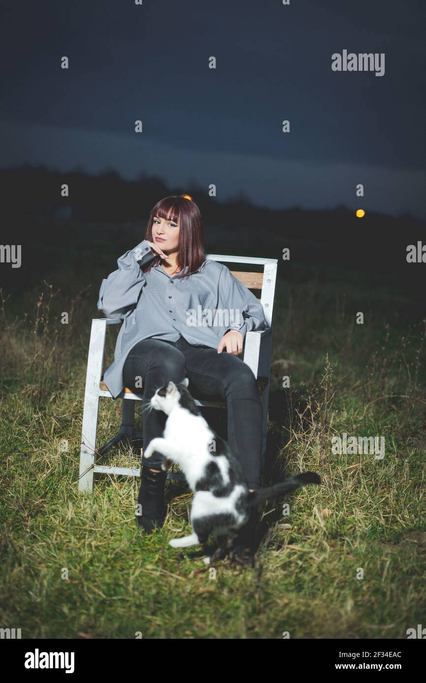 A vertical shot of a young caucasian fashionable female sitting ng on a chair in the field at night with a cat Stock Photo