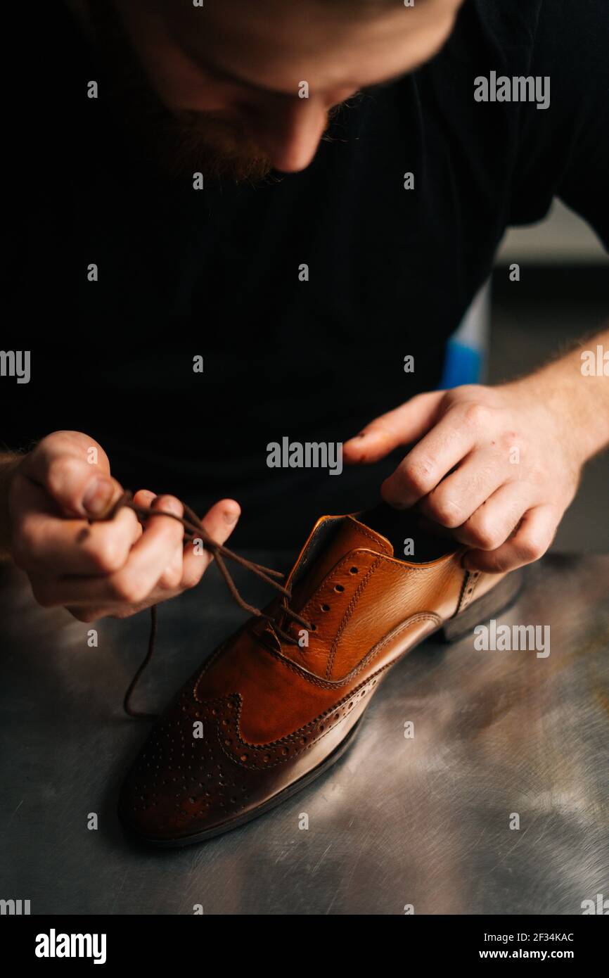 Close-up view of bearded male shoemaker tying laces on repaired and polished light brown leather shoes. Stock Photo