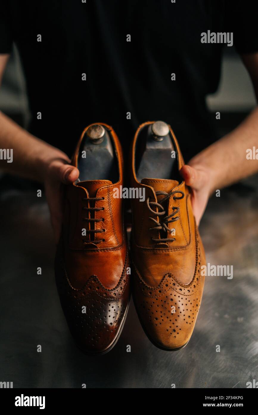 Close-up hands of male shoemaker holds old light brown leather shoe and repaired shiny shoes after restoration working. Stock Photo
