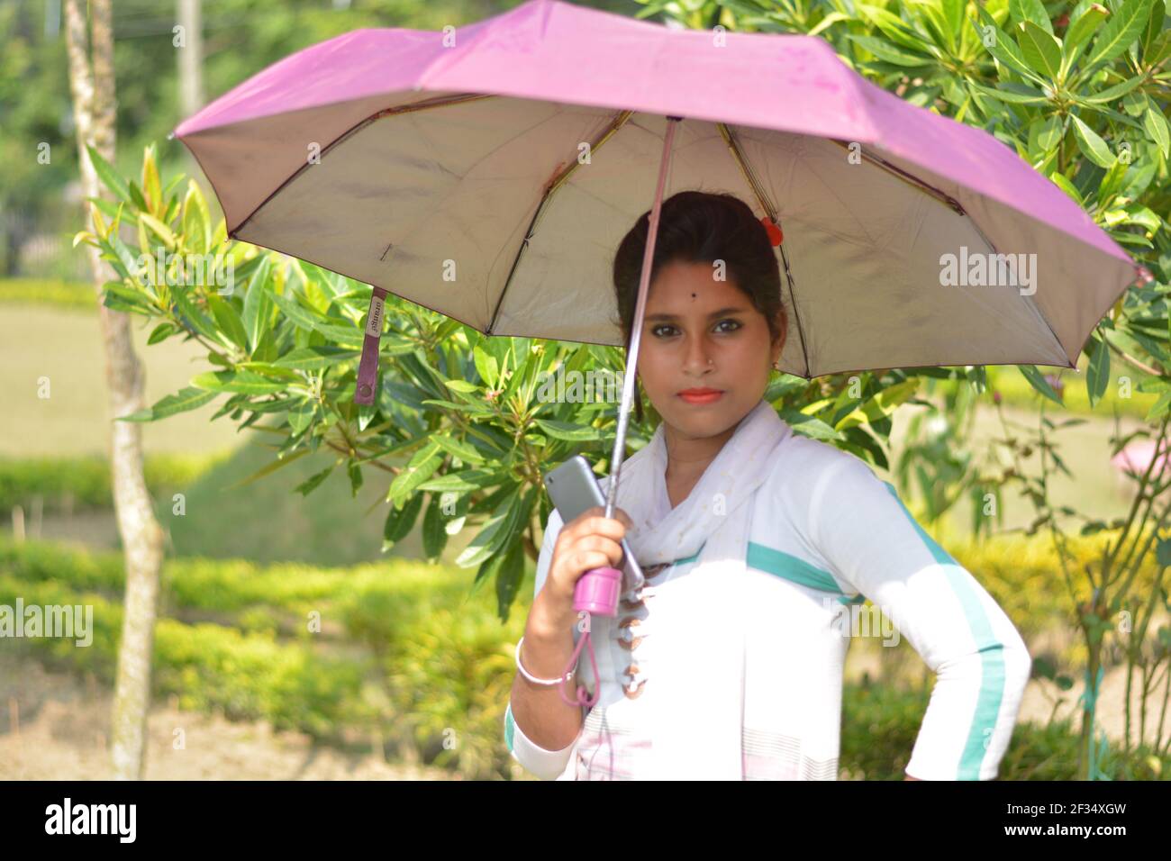 Close up of Indian Bengali teenage girl wearing white cotton salwar dhupatta holding mobile and umbrella on head,  golden nose pin and earring black Stock Photo
