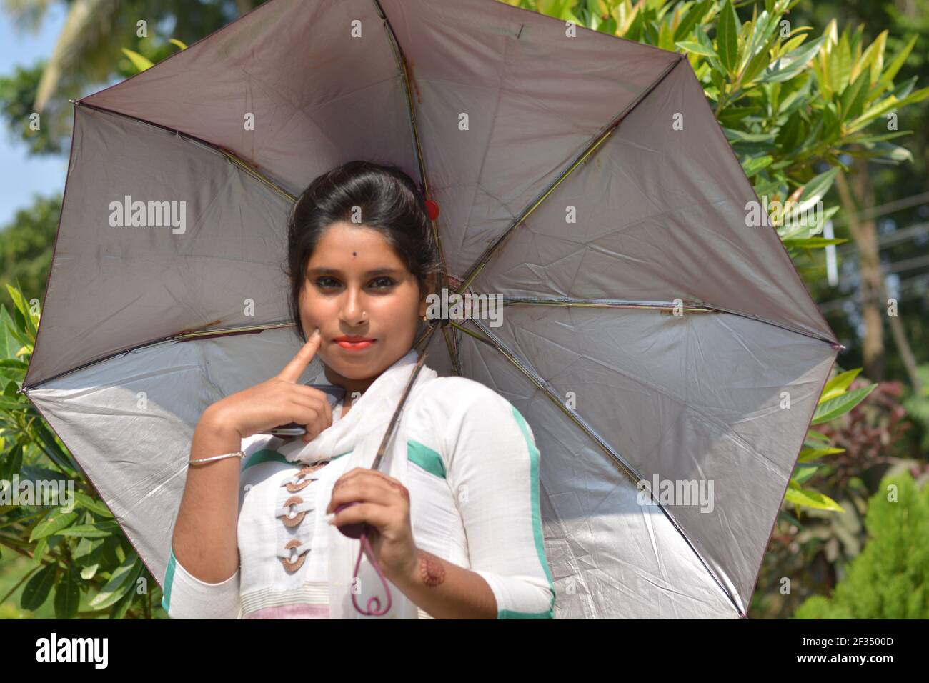 Close up of an Indian Bengali teenage girl wearing white cotton salwar black hair umbrella on shoulder finger on cheeks smiling, selective focusing Stock Photo