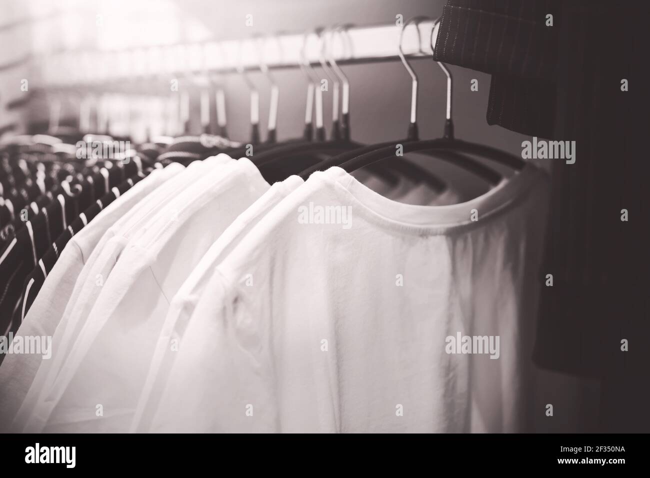 A black and white image of simple everyday T-shirts hanging on hangers in the wardrobe. Shopping in a clothing store. Stylish things. Stock Photo