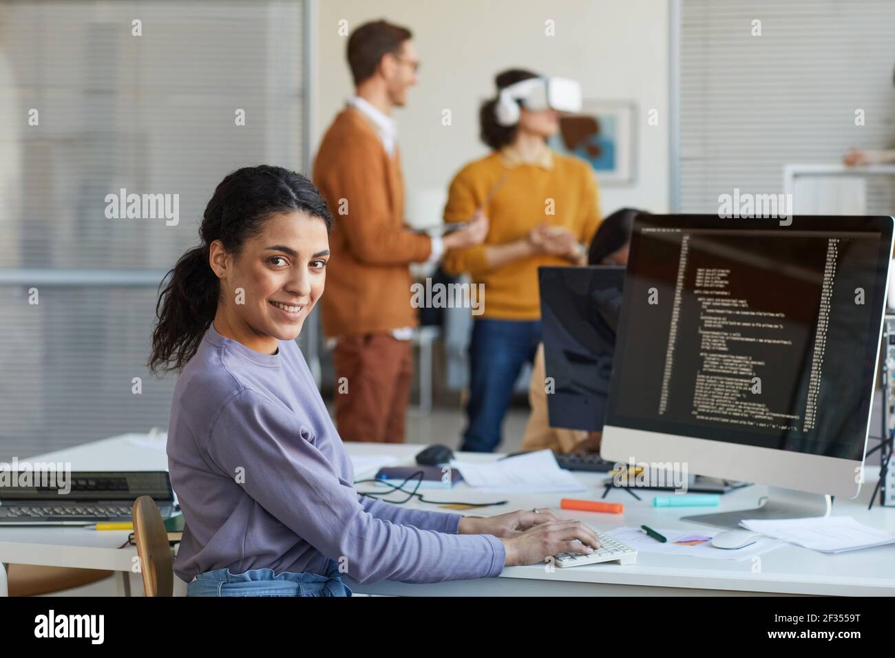 Portrait of female IT developer looking at camera and smiling while using computer with code on screen in software production studio, copy space Stock Photo