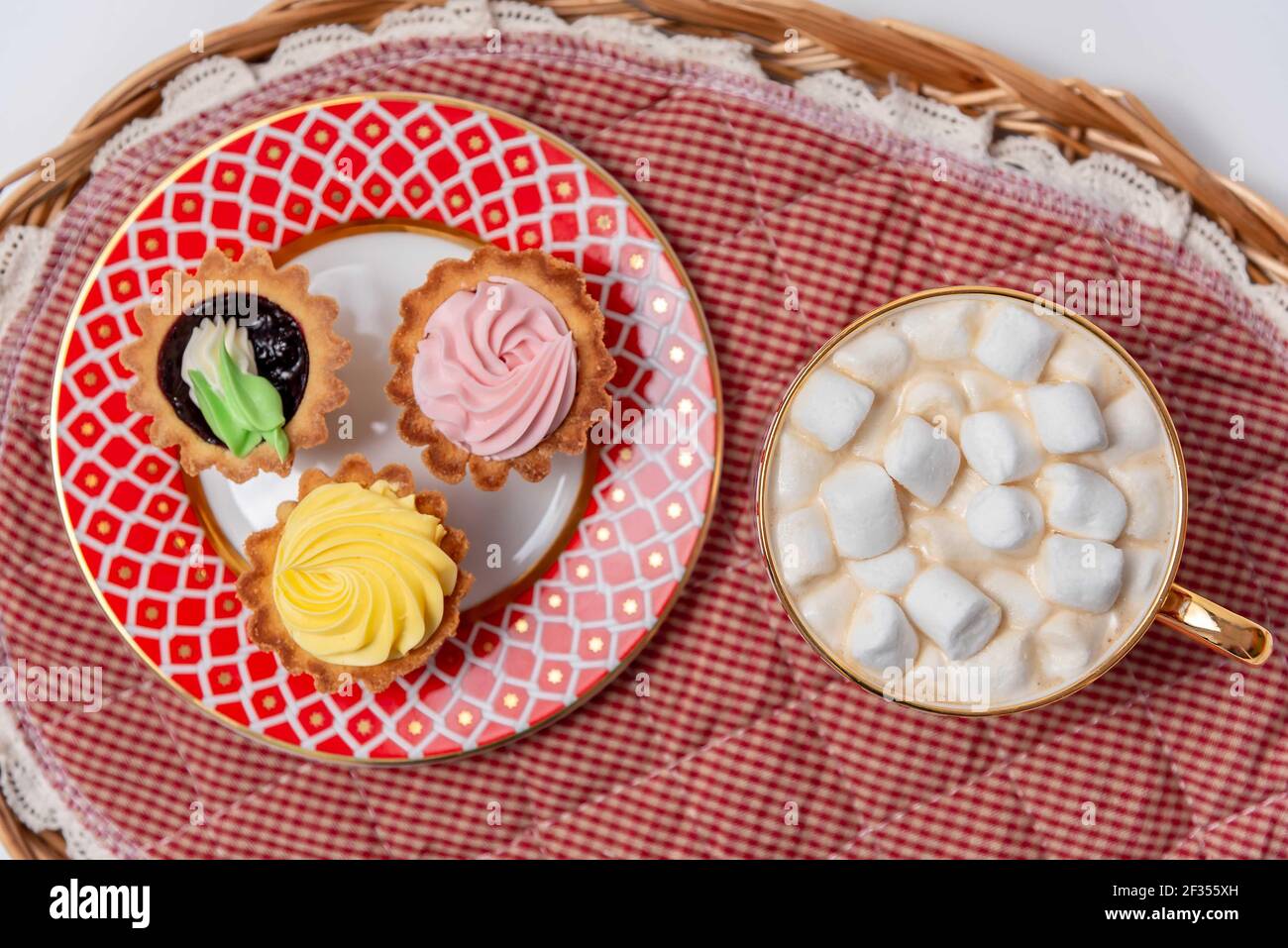 Tasty cupcakes and cup of cocoa with marshmallows on wicker tray, vanilla cupcakes with pink and yellow cream. Stock Photo