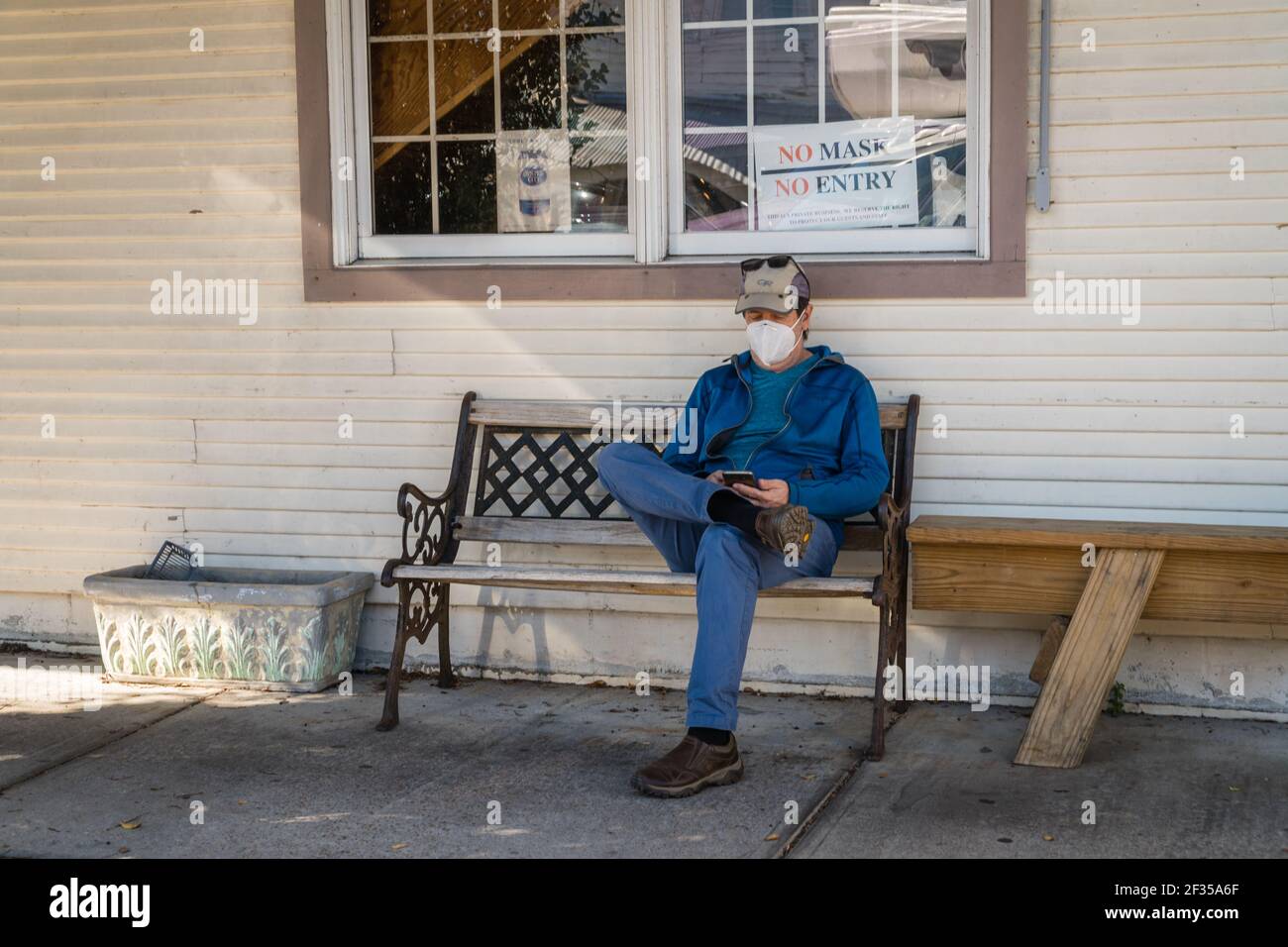 Man sitting on bench outside of restaurant wearing mask during the Covid-19 pandemic. Behind him is a sign reading 'no mask, no entry'. Stock Photo