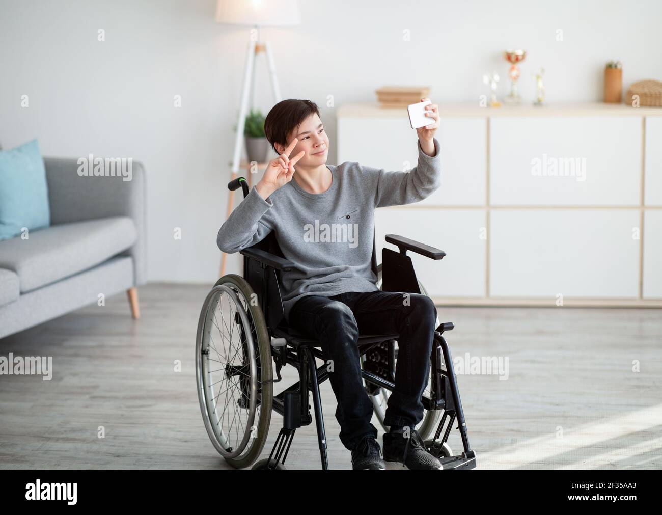 Happy disabled teenager in wheelchair taking selfie on smartphone, smiling at camera indoors Stock Photo