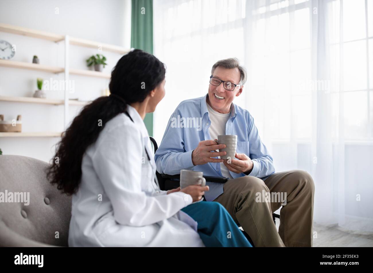 Happy disabled elderly patient and young doctor drinking tea together during medical visit at home Stock Photo