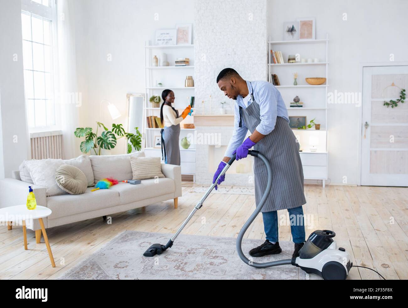 Full length of young black guy vacuuming floor while his wife dusting