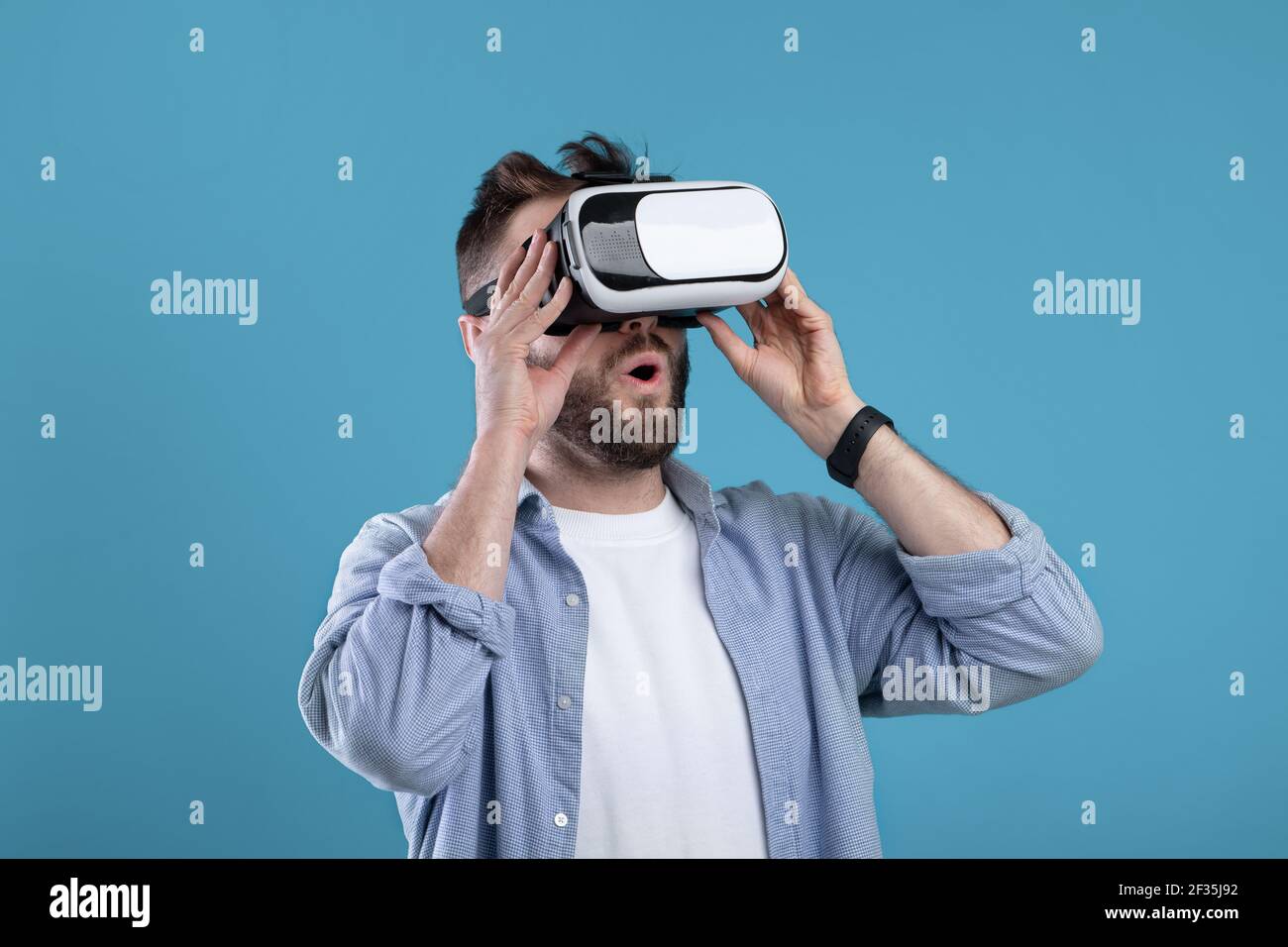 Cool millennial guy experiencing virtual reality in VR headset on blue studio background Stock Photo