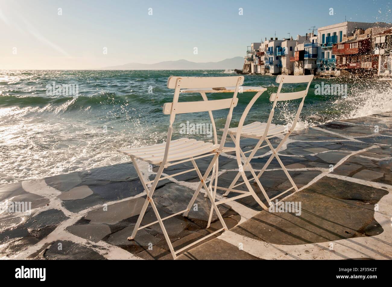 Two empty chairs on the waterfront in the island of Mykonos, Greece. In the background the splash of the waves and the characteristic Greek houses on Stock Photo
