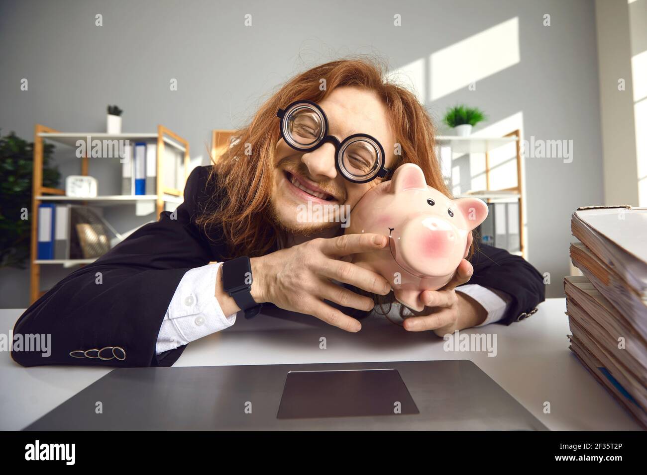 Funny guy sitting at office desk with laptop, hugging piggy bank and smiling happily Stock Photo