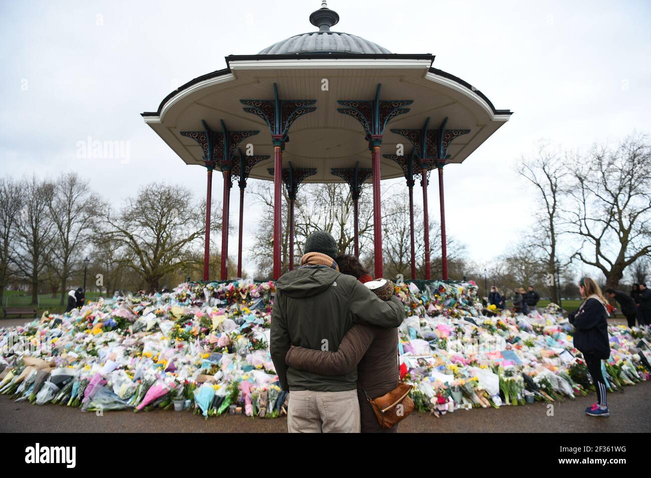 People stand next to floral tributes left at the band stand in Clapham Common, London, for murdered Sarah Everard. Serving police constable Wayne Couzens, 48, appeared in court on Saturday charged with kidnapping and murdering the 33-year-old marketing executive, who went missing while walking home from a friend's flat in south London on March 3. Picture date: Monday March 15, 2021. Stock Photo