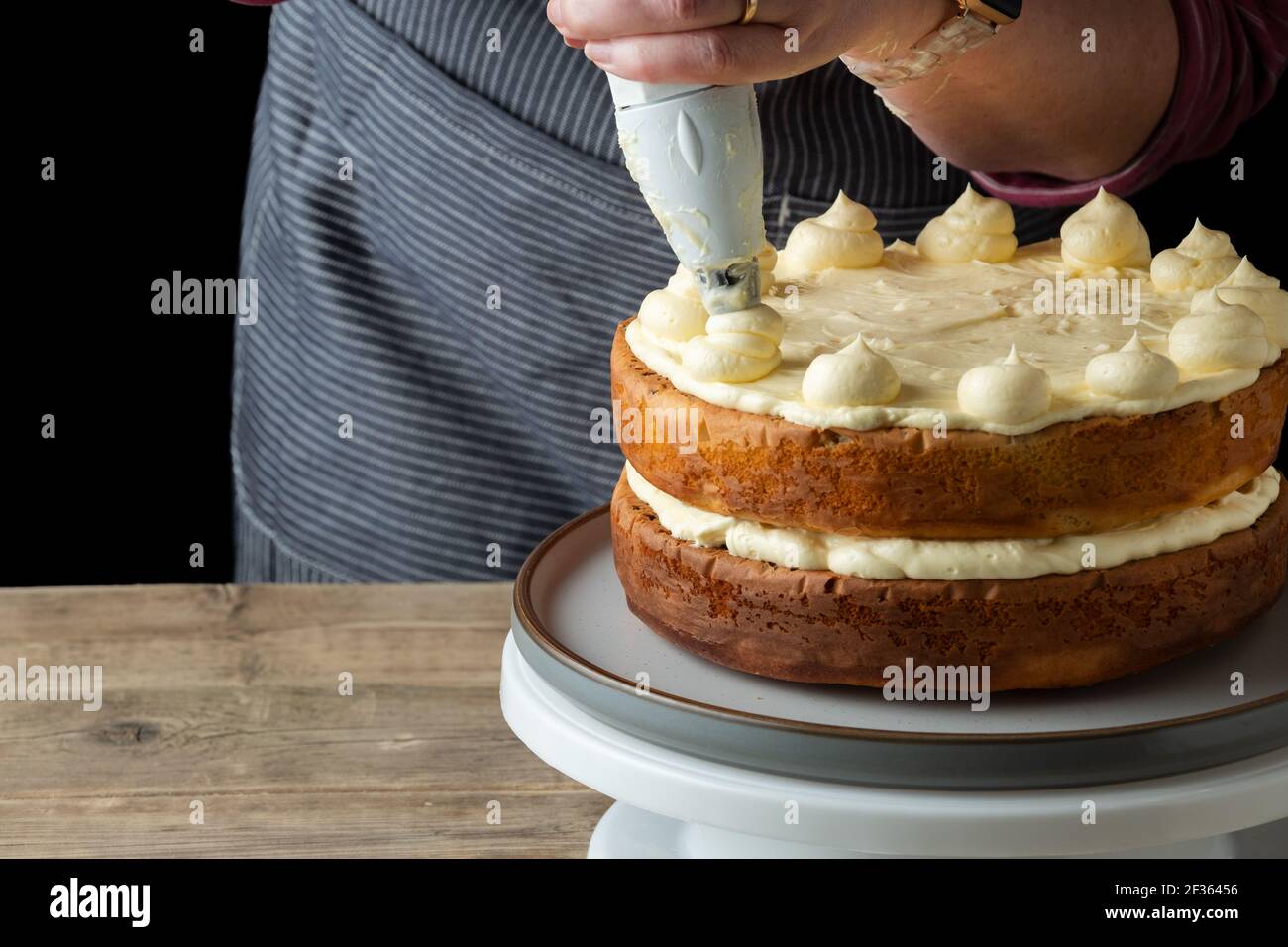 A baker adds butter cream to the top of a freshly home baked cake. The baker, a female is skillfully adding the butter cream using a piping bag Stock Photo