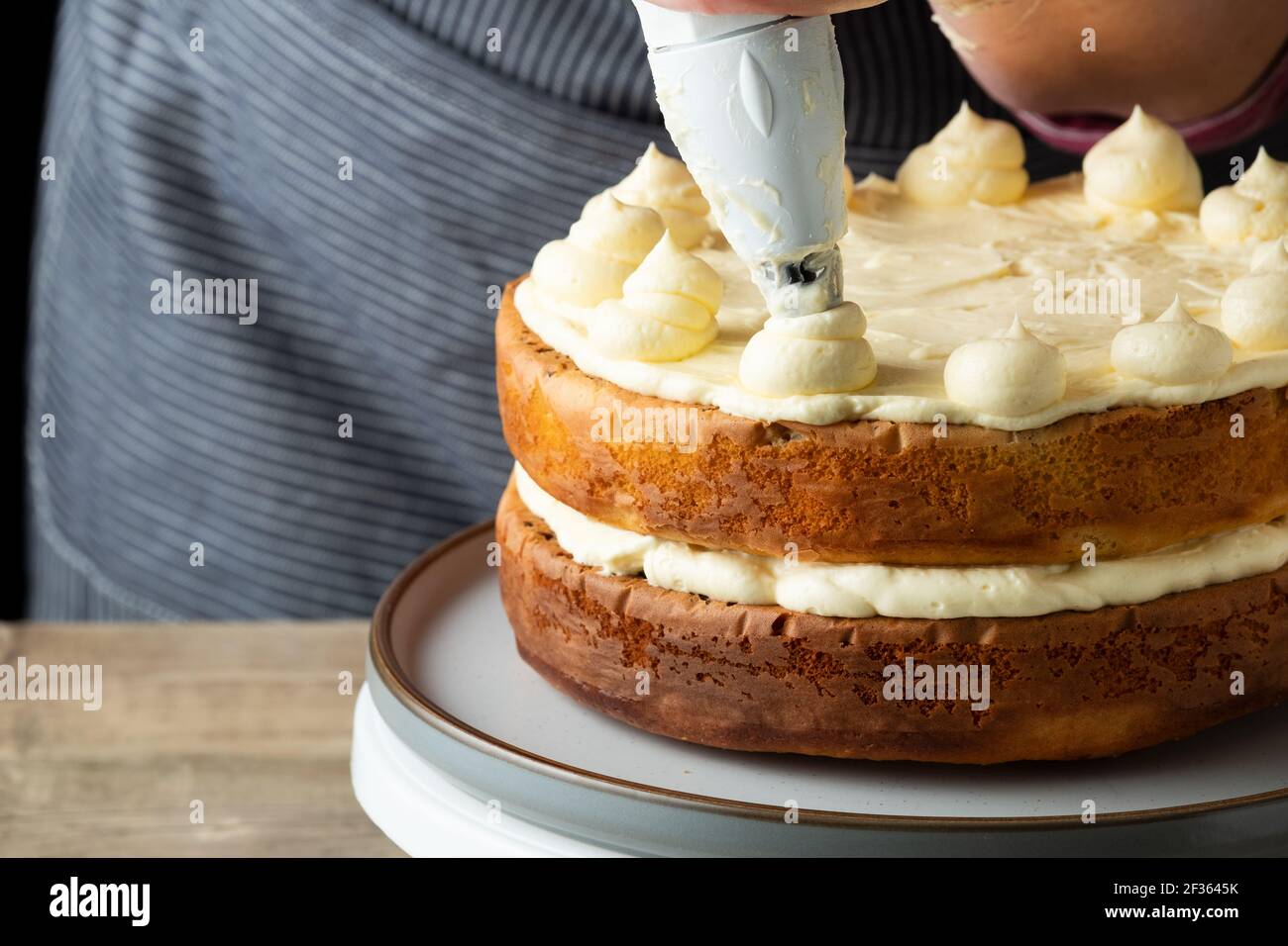 A baker adds butter cream to the top of a freshly home baked cake. The baker, a female is skilfully adding the butter cream using a piping bag Stock Photo