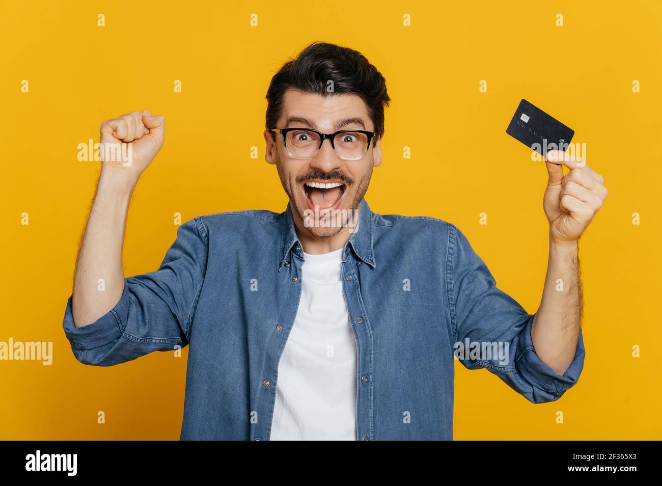Young excited positive caucasian man holds credit card, rejoices in success, victory, profit, standing on isolated orange background happily raises his fist and smiling Stock Photo