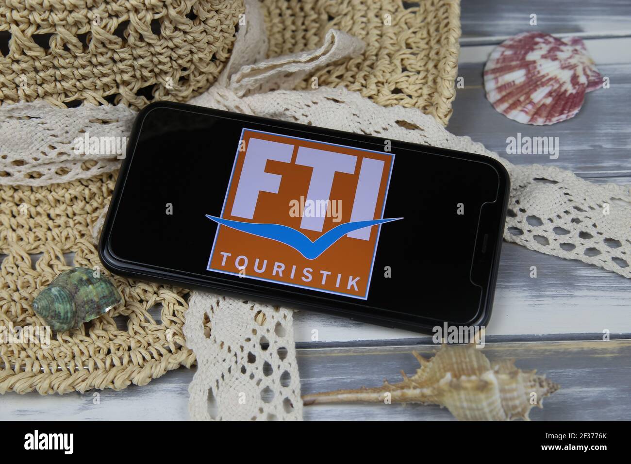 Viersen, Germany - March 1. 2021: Closeup of smartphone with logo lettering of fti touristik group travel agency with sun hat and shells on wood table Stock Photo