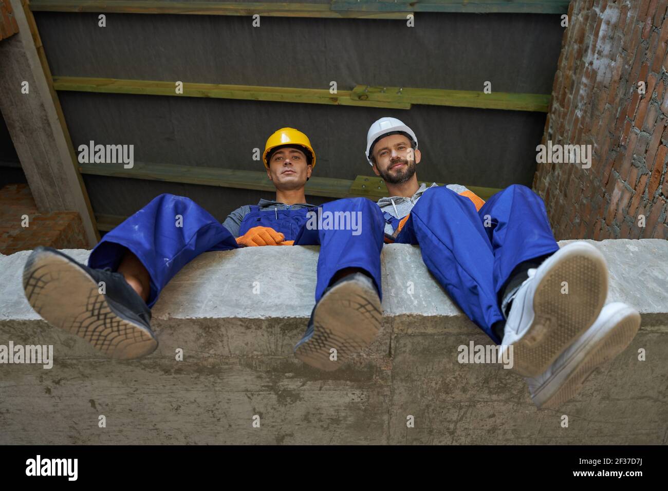 Two attractive young builders in blue overalls and hard hats looking down at camera, sitting on the concrete floor, resting while working on house Stock Photo