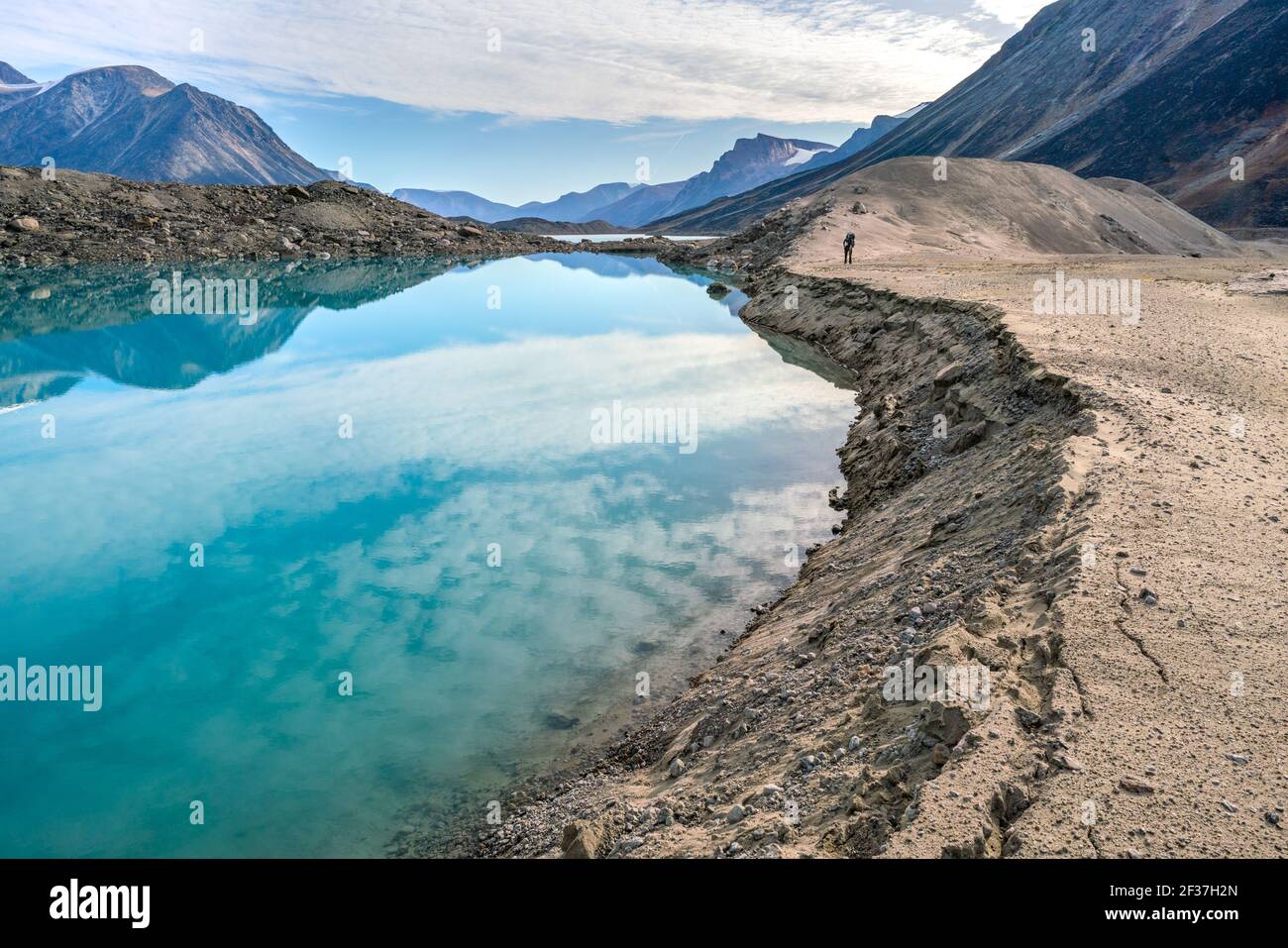 Hiker walking by a lake through remote arctic valley on a partly cloudy summer day. Dramatic arctic landscape of Akshayuk Pass, Baffin Island, Canada. Stock Photo