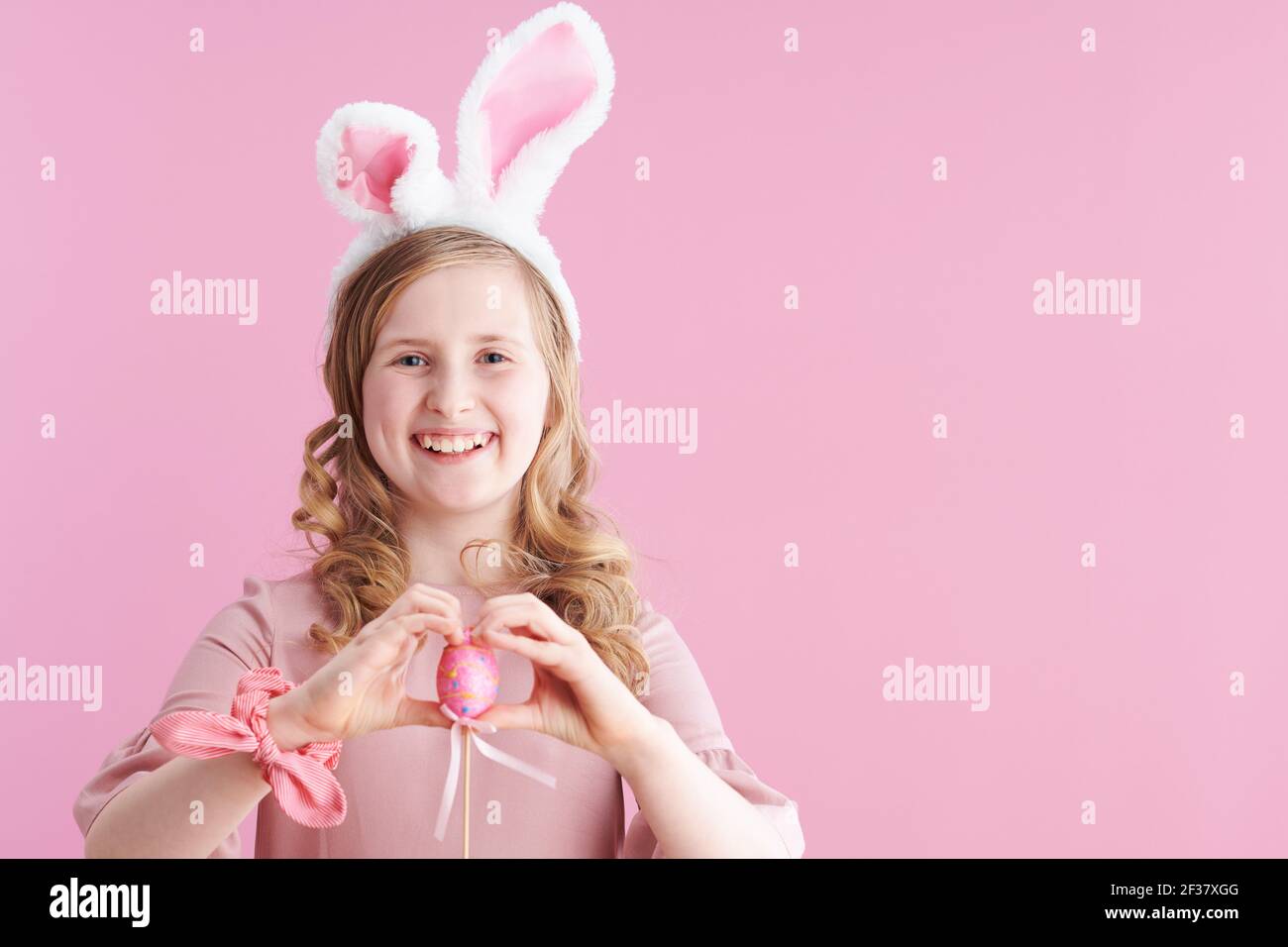 Portrait of happy stylish child in pink dress with bunny ears and easter egg against pink background. Stock Photo