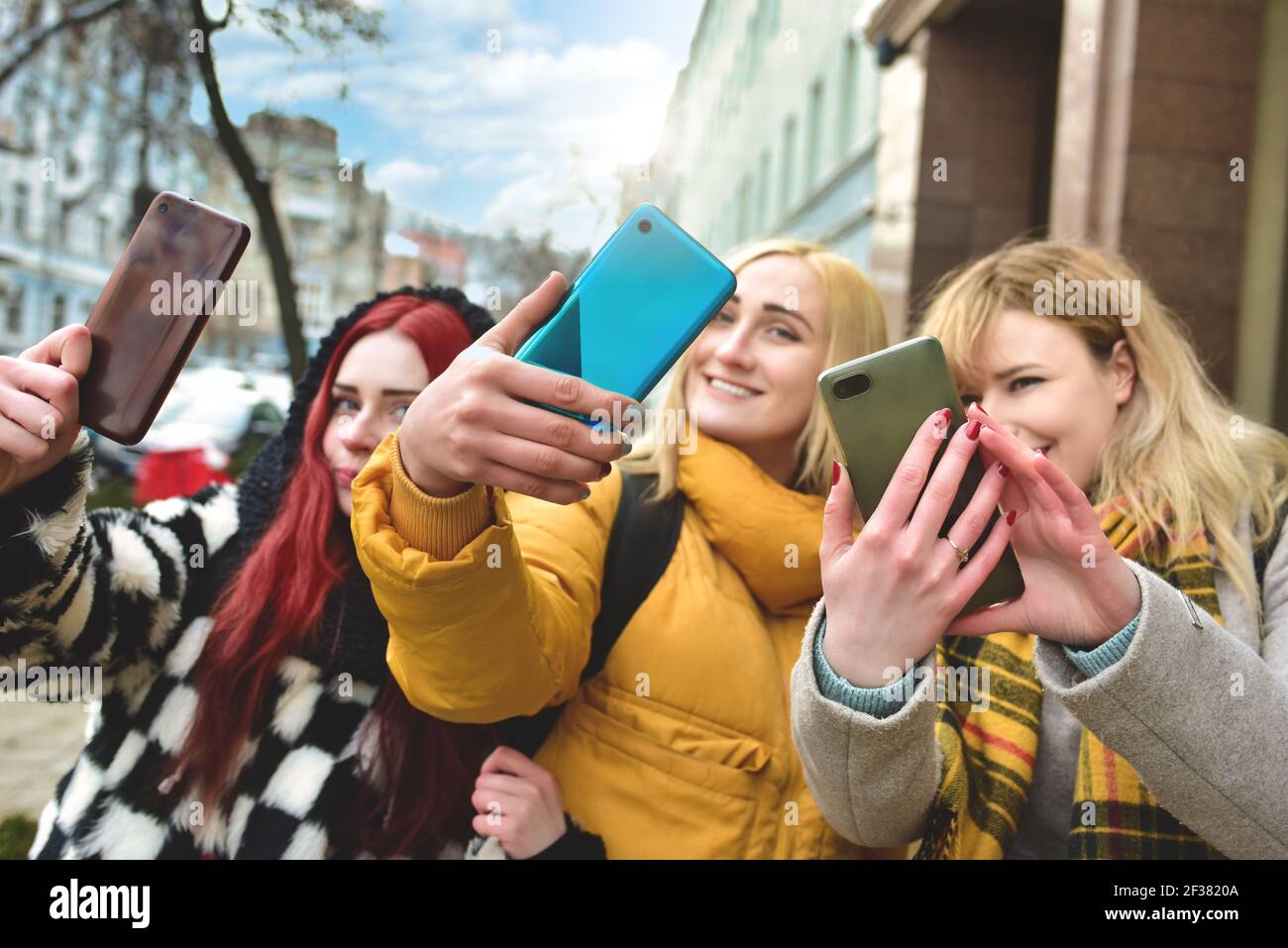 Three beautiful smiling young women walk in the city, using phone outdoors make selfies Stock Photo