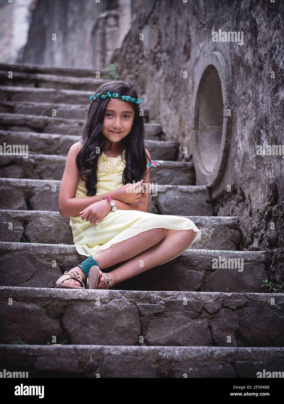Little girl in yellow dress sitting on the steps of a park with a wreath of flowers on her head smiles at the camera Stock Photo