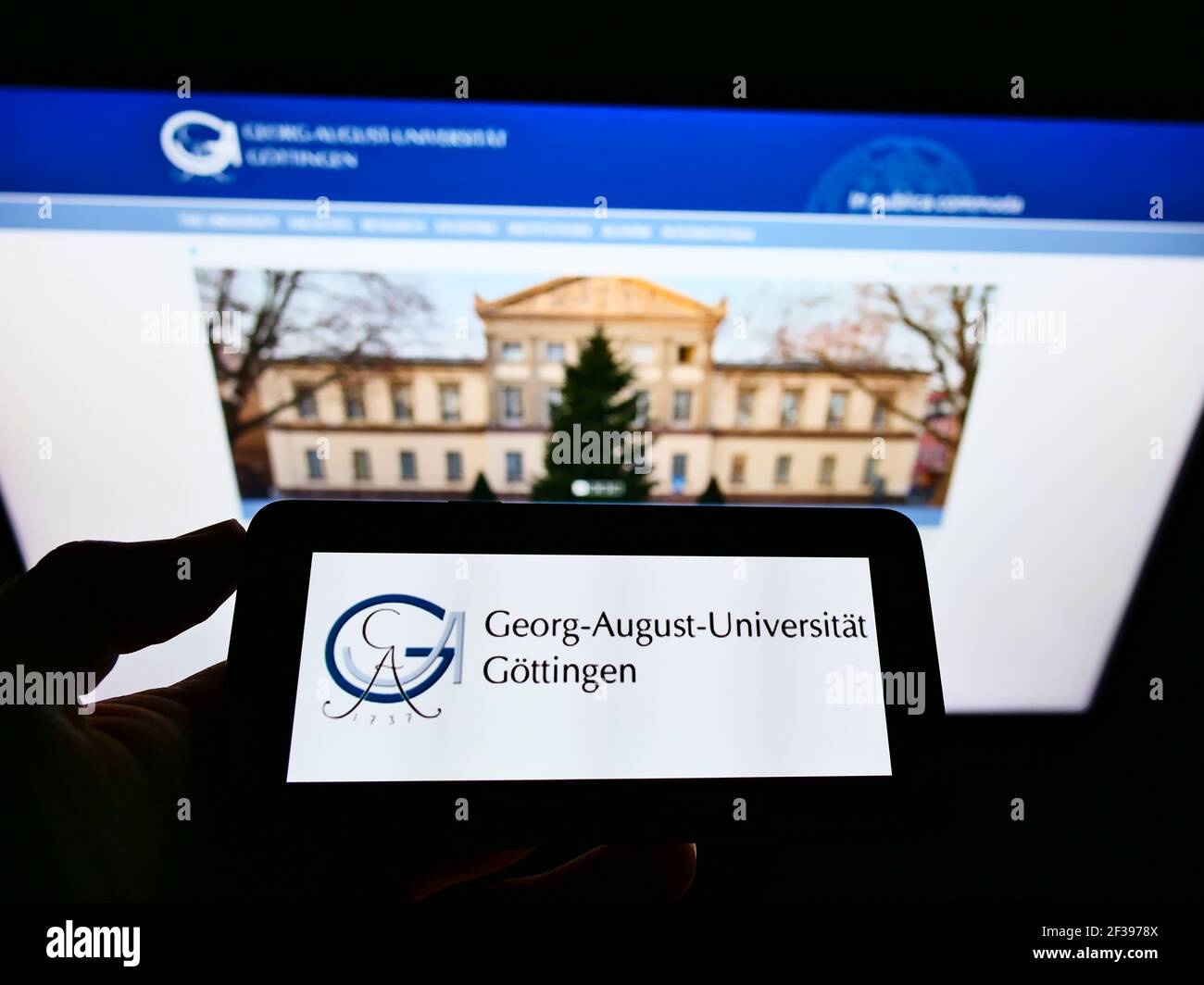 Person holding mobile phone with logo of German education institution University of Göttingen on screen in front of web page. Focus on phone display. Stock Photo