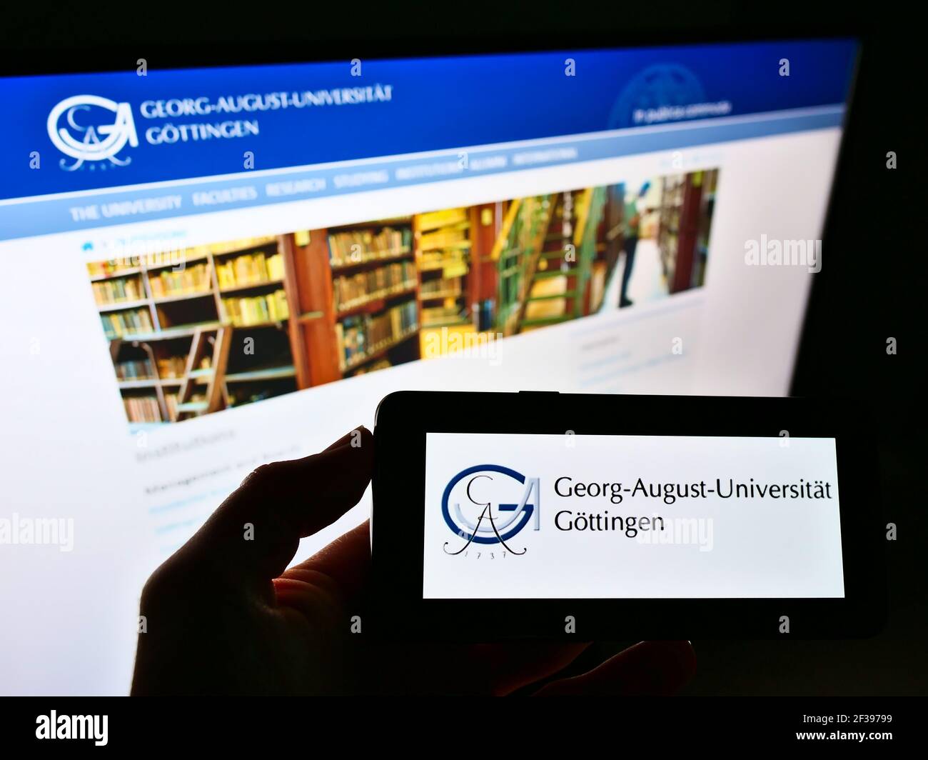 Person holding smartphone with logo of German university Georg-August-Universität Göttingen on screen in front of website. Focus on phone display. Stock Photo