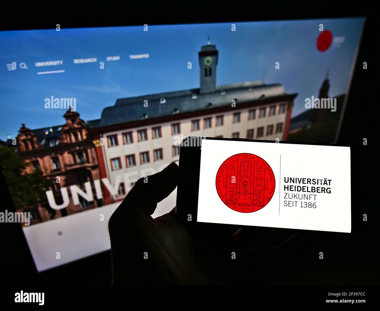 Person holding cellphone with logo of German university Ruprecht-Karls-Universität Heidelberg on screen in front of web page. Focus on phone display. Stock Photo
