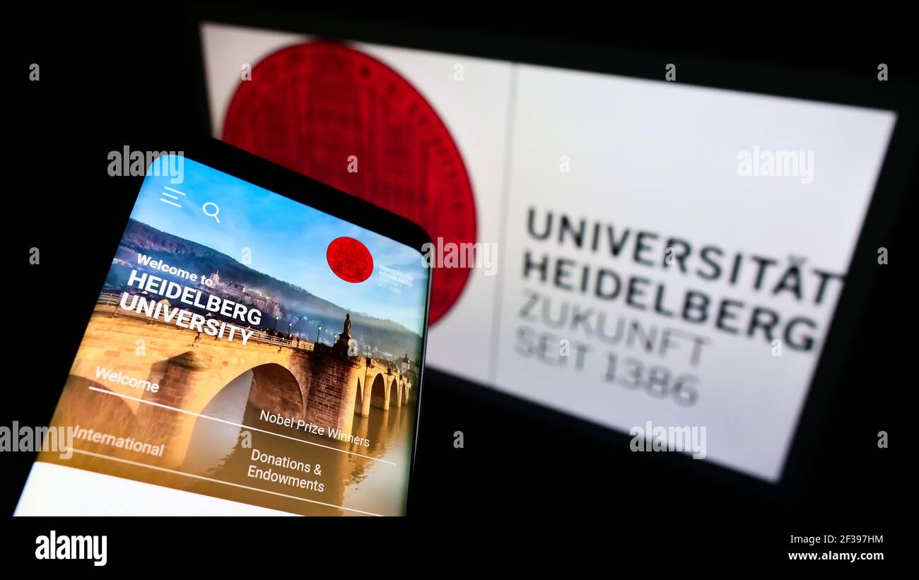 Person holding mobile phone with web page of German Heidelberg University on screen in front of logo. Focus on top-left of phone display. Stock Photo