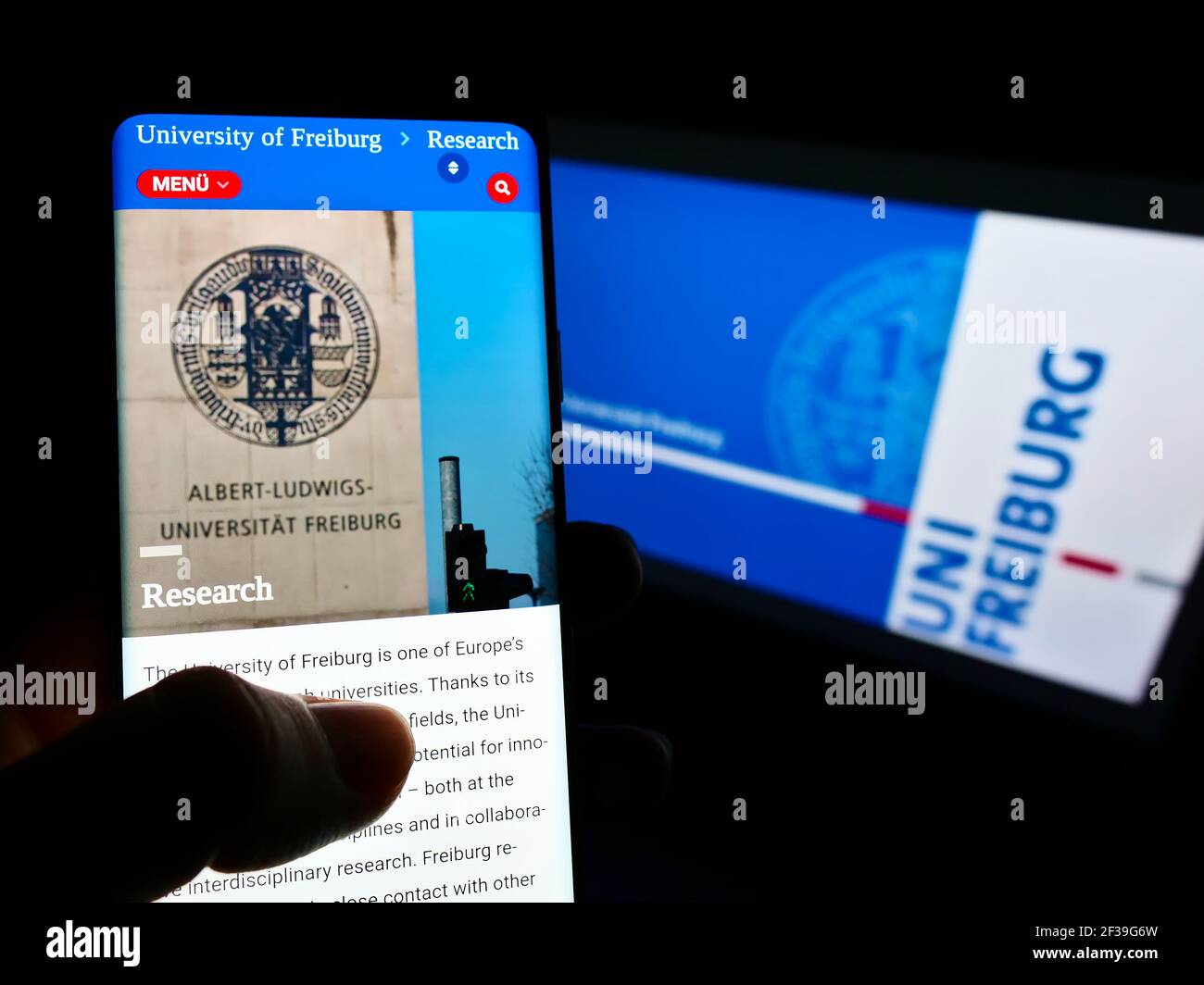 Person holding mobile phone with webpage of German University of Freiburg on screen in front of logo. Focus on center of cellphone display. Stock Photo