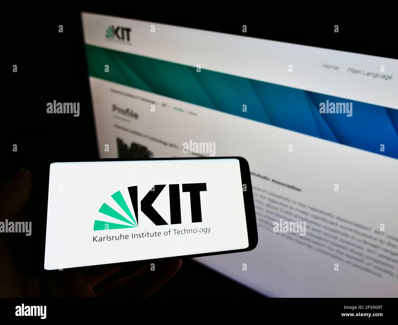 Person holding mobile phone with logo of German Karlsruhe Institute of Technology (KIT) on screen in front of web page. Focus on cellphone display. Stock Photo