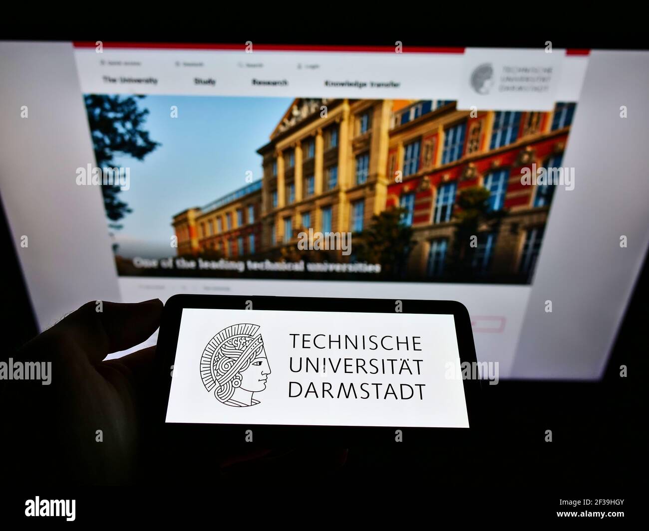 Person holding smartphone with logo of German university Technische Universität Darmstadt on screen in front of website. Focus on phone display. Stock Photo