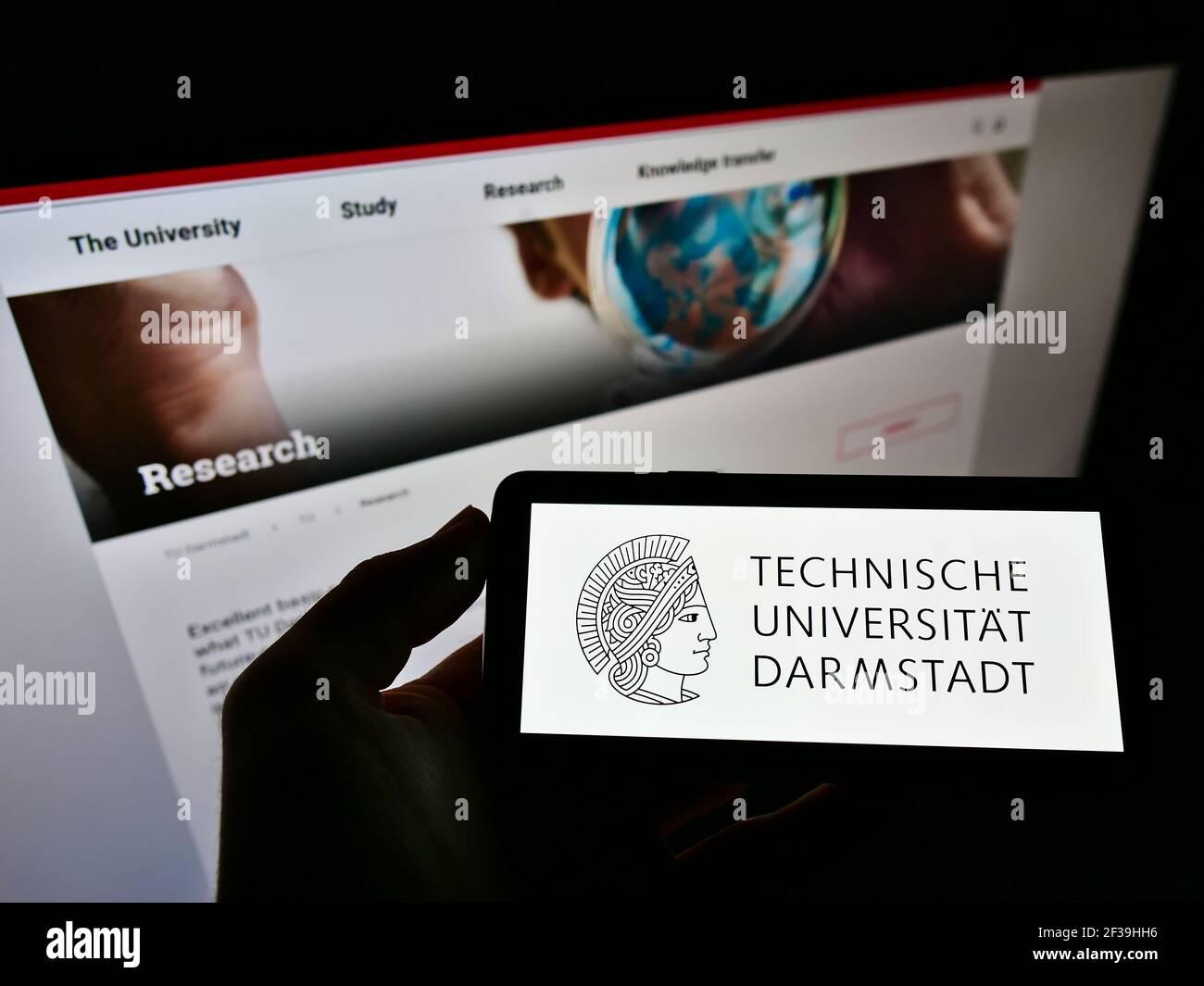 Person holding mobile phone with logo of German Technical University of Darmstadt on screen in front of web page. Focus on cellphone display. Stock Photo