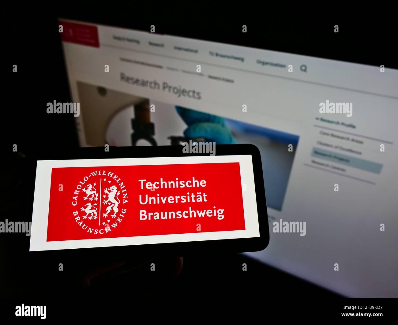Person holding smartphone with logo of German university Technische Universität Braunschweig on screen in front of webpage. Focus on phone display. Stock Photo
