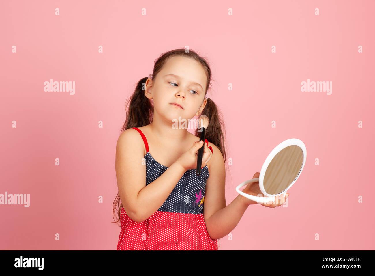 a European girl with ponytails and a dress, who powders her cheek with a brush, looks into a white plastic mirror, isolated on a pink background Stock Photo