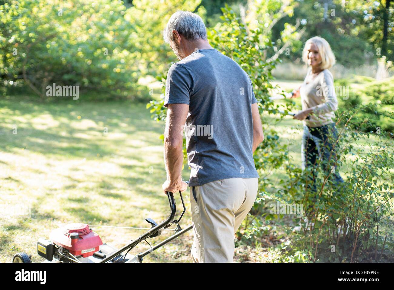 Mature man mowing lawn in backyard while his wife working in background Stock Photo