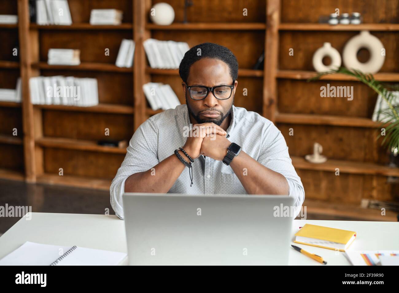 Concentrated black business man pursed lips sitting in front of a laptop, put his chin on hands, working on the computer with documents on the desk, thinking about future steps, plans, looks at screen Stock Photo