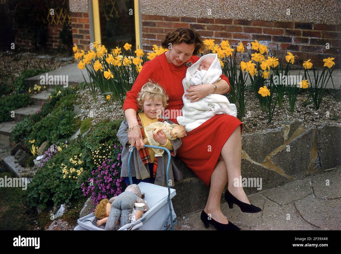 1960s, historical, springtime and daffodils are out, as a mother sitting outside at a rocky garden, holding her new baby in her left arm and holding the hand of her young daughter, who is carrying a doll and her little pram filled with soft toys, England, UK. Stock Photo