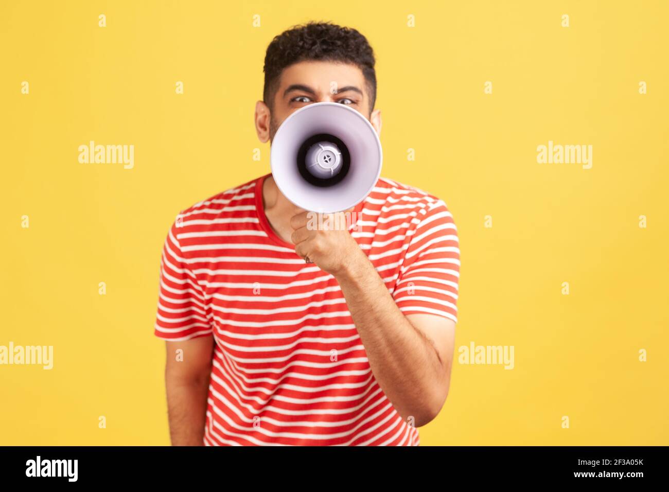 Annoyed angry man in striped t-shirt holding megaphone near mouth loudly speaking, screaming, making announcement, paying attention at social problems Stock Photo