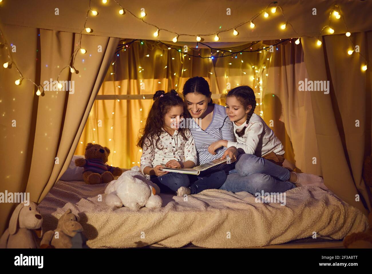 Happy mother and children sitting in playroom tent and reading book of fairy tales Stock Photo