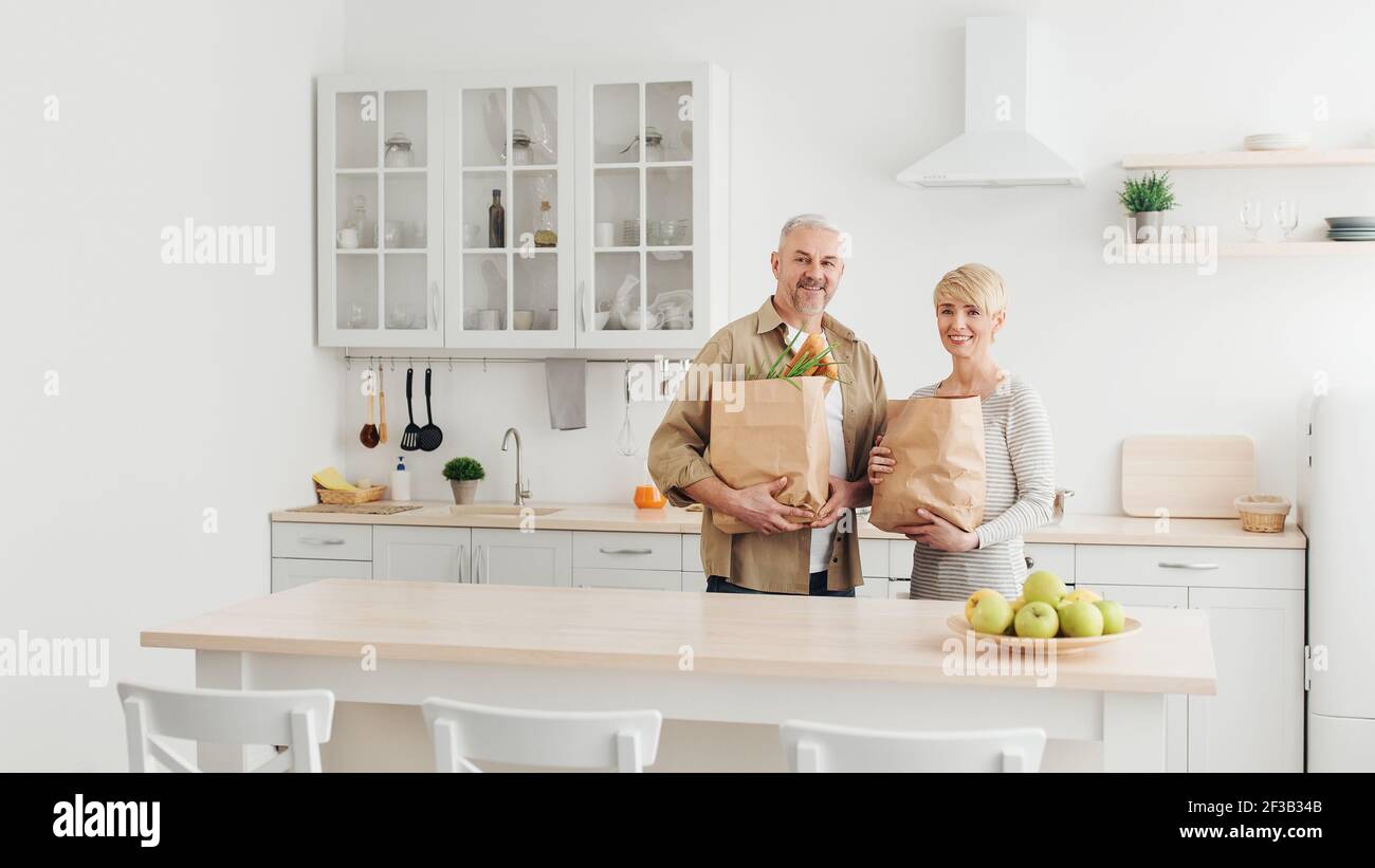 Happy married couple coming home from shopping and unpacking paper bags with groceries in kitchen together Stock Photo
