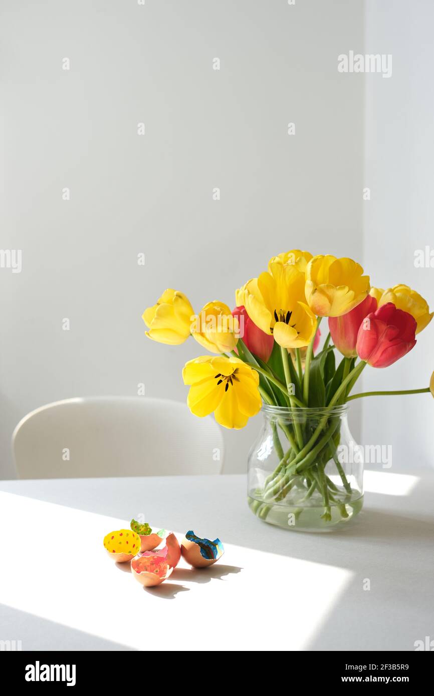 Children's craft of painted egg shells on a white table with a vase filled with yellow and red tulips in an otherwise white space Stock Photo