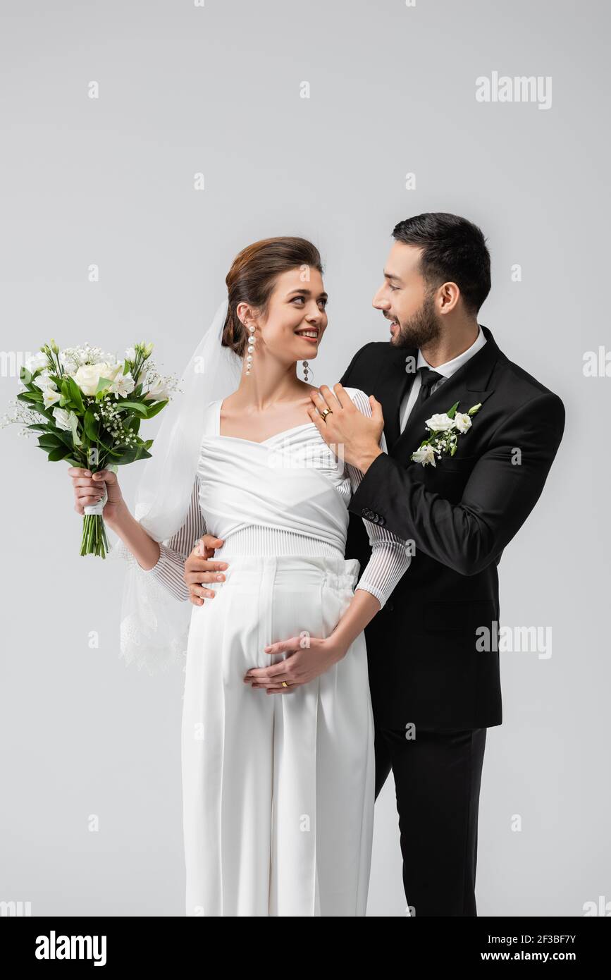 Muslim groom embracing pregnant bride with wedding bouquet isolated on grey Stock Photo