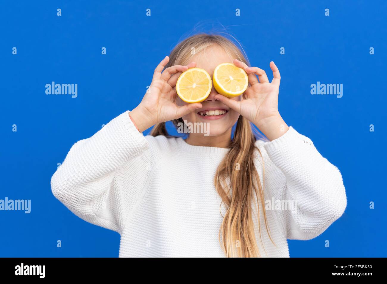 Overjoyed blond haired girl in white sweater smiling while covering eyes with bright lemon slices isolated on blue background. High quality photo Stock Photo