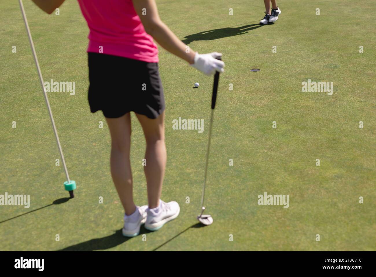 Caucasian woman playing golf holding club and flag while other player putts for the hole Stock Photo