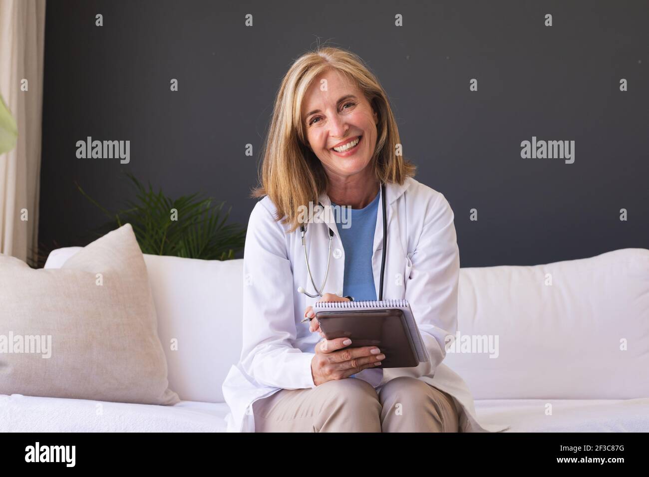 Caucasian senior female doctor sitting on sofa smiling giving video call consultation Stock Photo