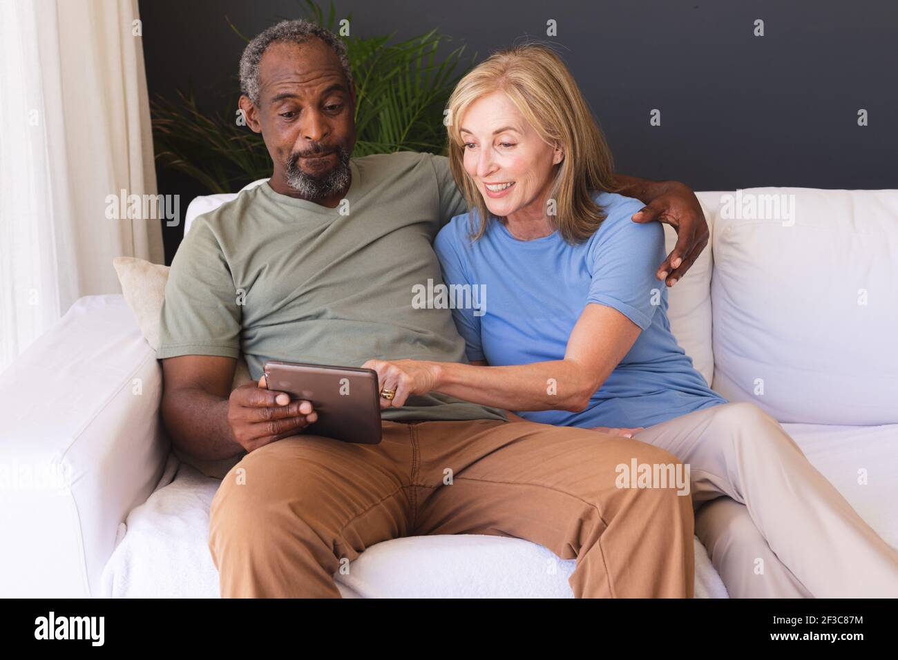 Diverse senior couple sitting on couch and using digital tablet Stock Photo