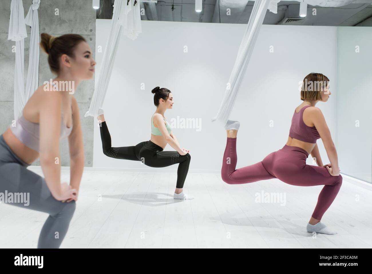 sportive women stretching with fly yoga hammocks in fitness center, blurred foreground Stock Photo