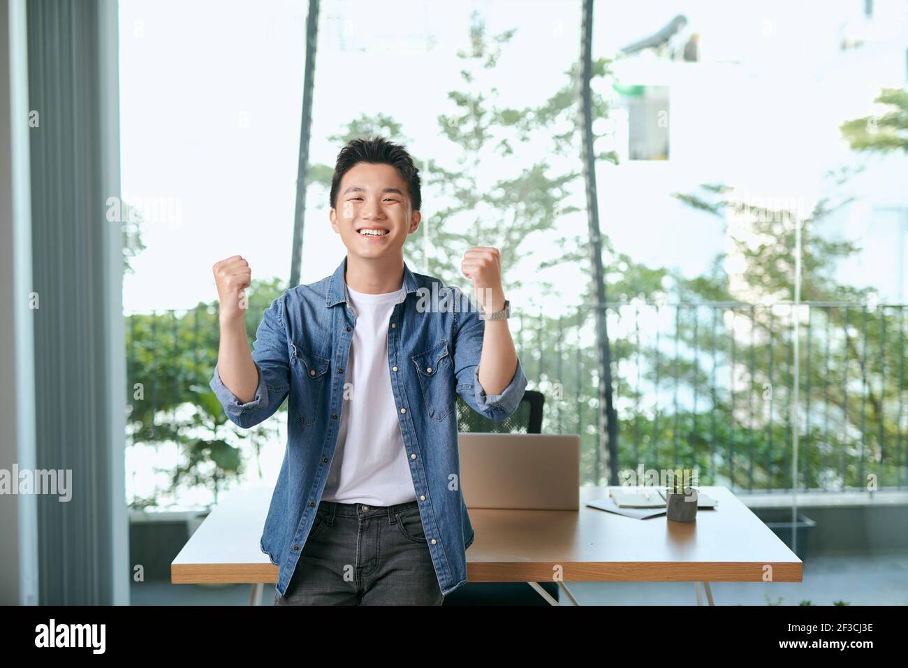 Everyday winner. Cheerful young man in casual wear keeping arms raised and looking happy while sitting at the desk in office Stock Photo