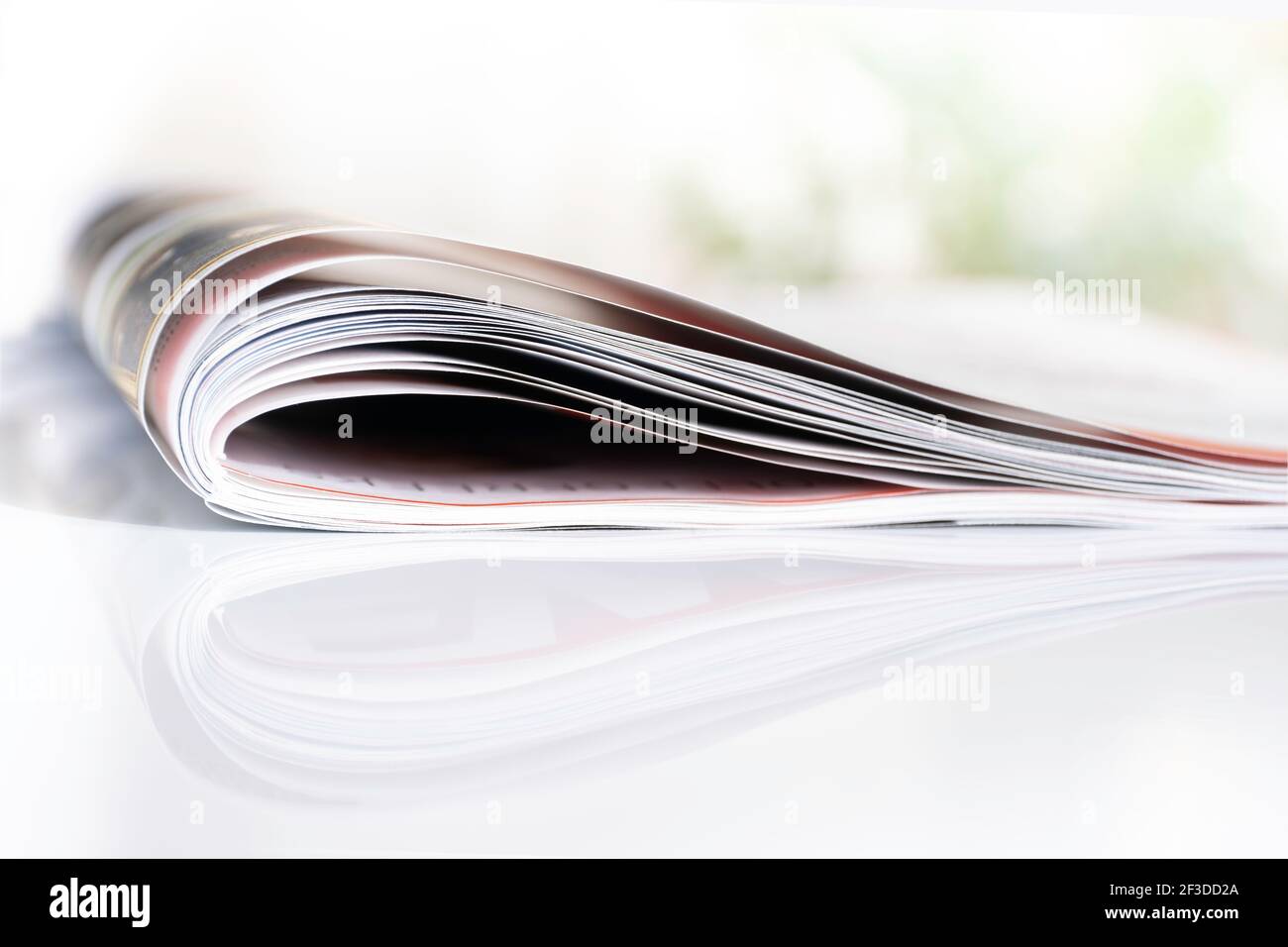 Folded magazine reflected on a white surface with shallow depth of field. Blurred background of green bushes Stock Photo